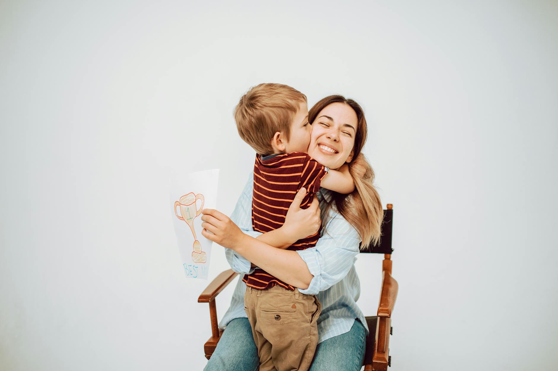 A joyful mother holding her son, who is giving her a kiss, while he displays a colorful drawing of a trophy that says 'Best Mom'. They are sitting together against a plain background.